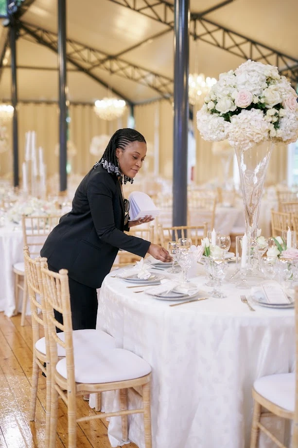 Une wedding planner en tailleur noir ajuste le couvert d’une table ronde aux nappes blanches, sous une verrière ornée de lustres, près d’un grand bouquet d’hortensias et de roses