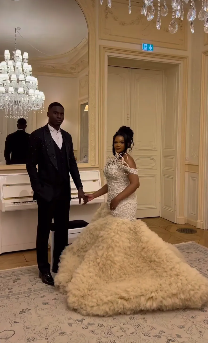 Film : un couple de mariés, main dans la main, dans un salon au lustre de cristal, la robe de la mariée en plumes champagne déployée