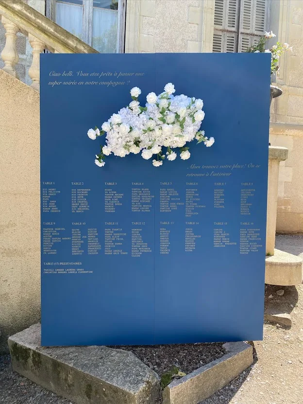 Plan de table sur panneau bleu, orné d’une composition de fleurs blanches, devant une façade en pierre