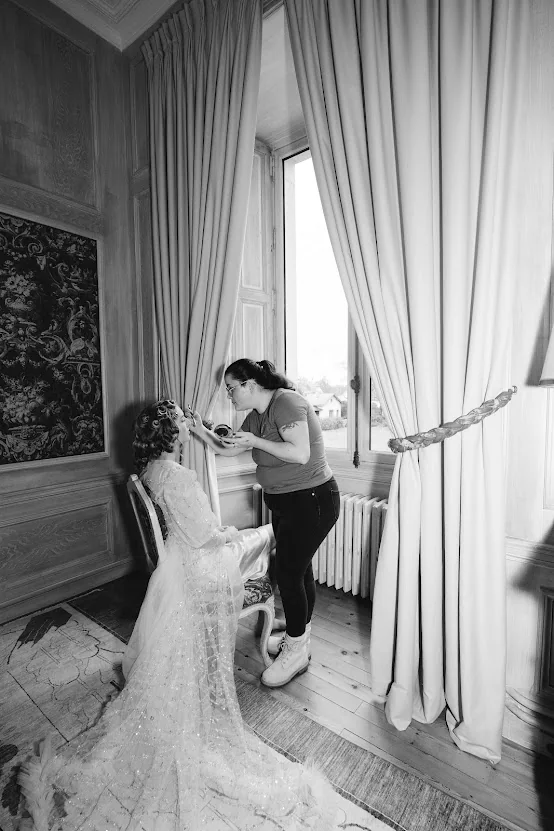 Préparatifs de la mariée en noir et blanc : une maquilleuse la coiffe près d’une grande fenêtre, dans une chambre de château