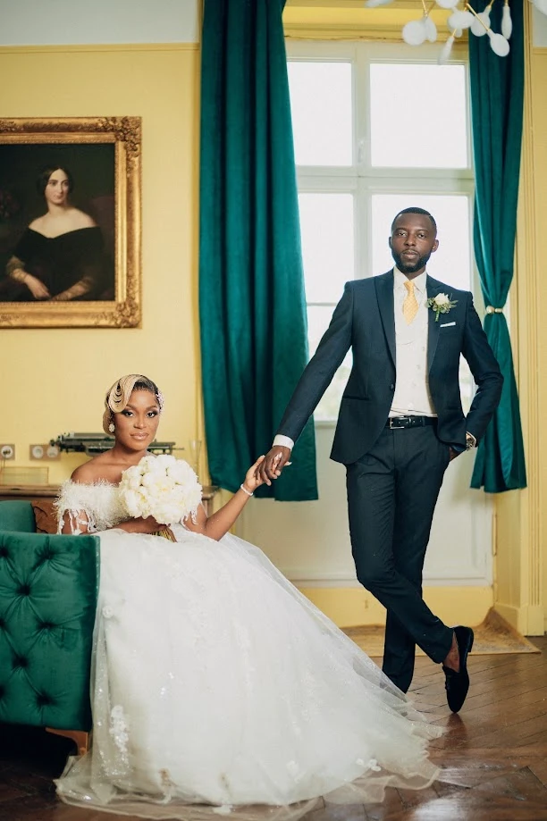 Dans un salon de château jaune aux rideaux vert émeraude, la mariée est assise sur un canapé en velours vert, un bouquet blanc sur les genoux ; le marié, debout, lui tient la main