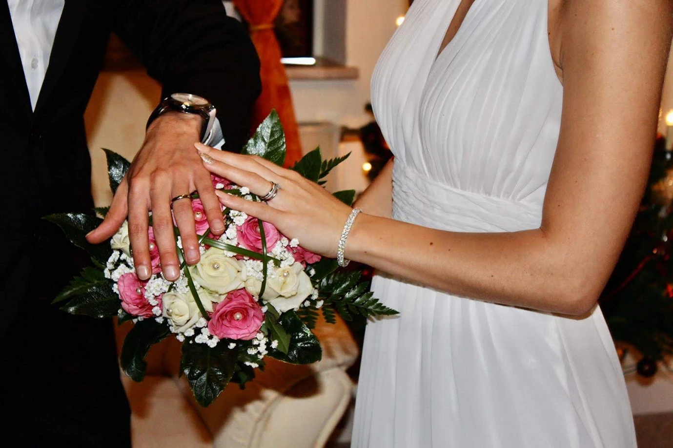 Les mains des mariés posées l’une sur l’autre sur un bouquet de roses roses et blanches, alliances aux doigts : le marié en costume sombre, la mariée en robe blanche plissée et bracelet de cristaux