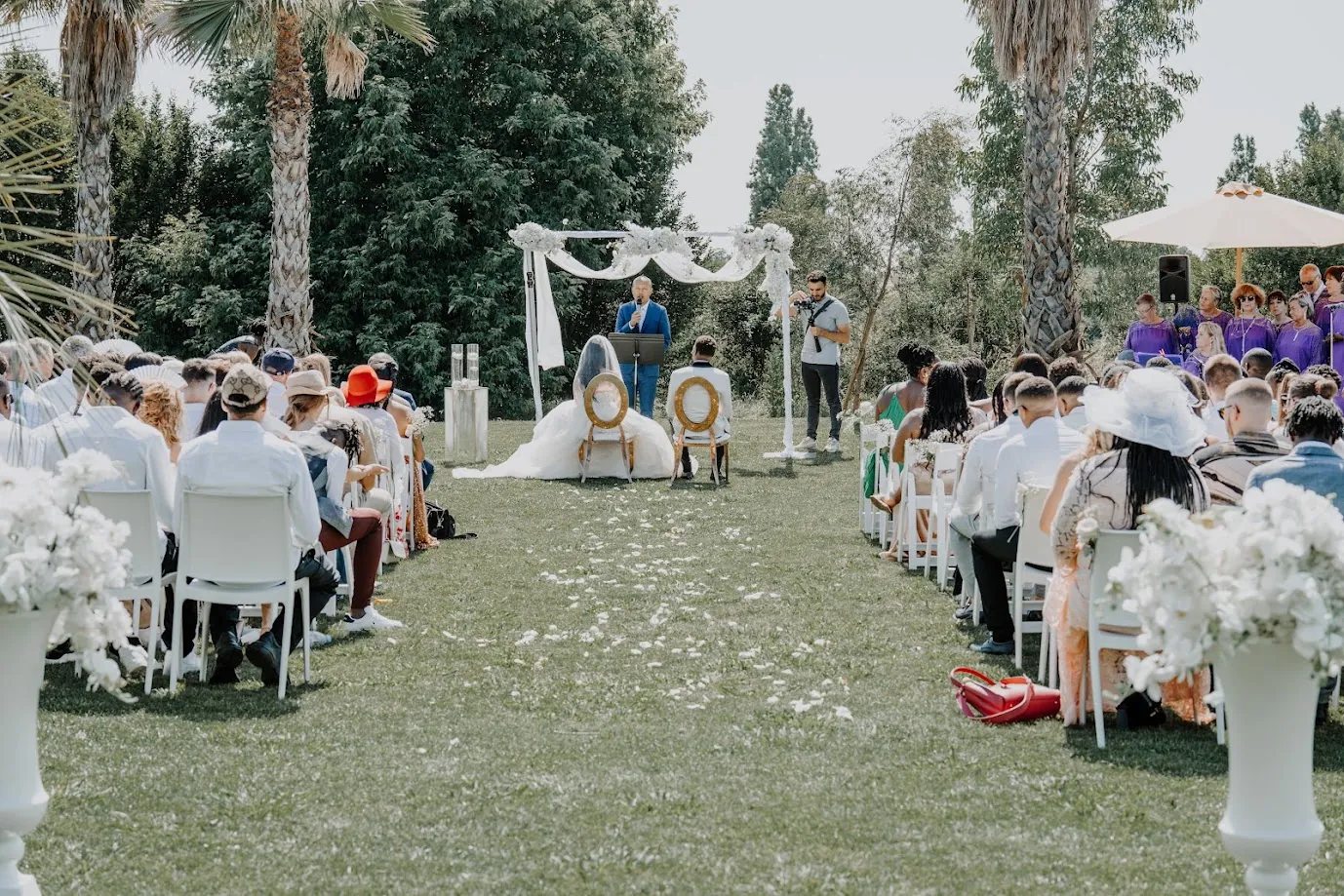 Cérémonie laïque en plein air : les mariés assis face à l’officiant sous une arche de voiles et de fleurs blanches, entourés de leurs invités et d’une chorale