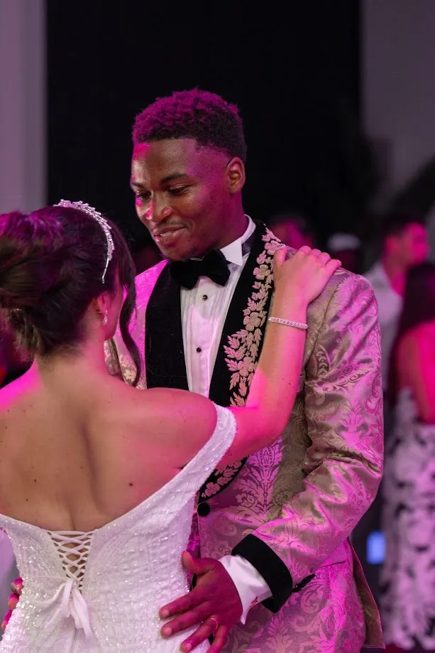 Pendant la soirée, la mariée, de dos, et le marié, en veste brodée de feuillages dorés, dansent dans une lumière rose