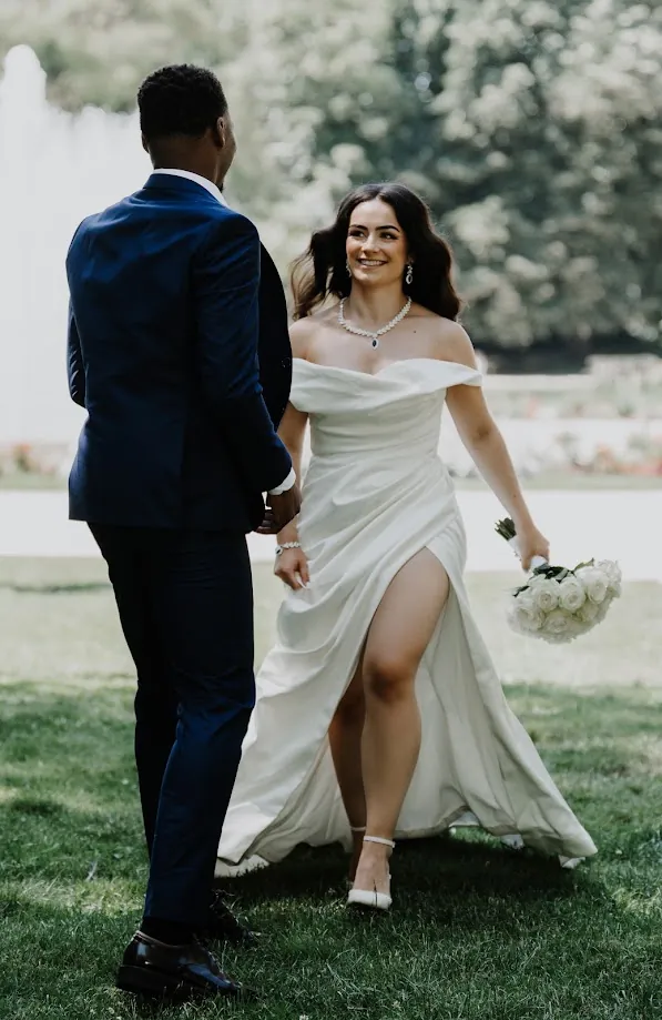 Dans un parc, la mariée, robe fendue et bouquet blanc à la main, s’avance vers le marié en costume bleu nuit