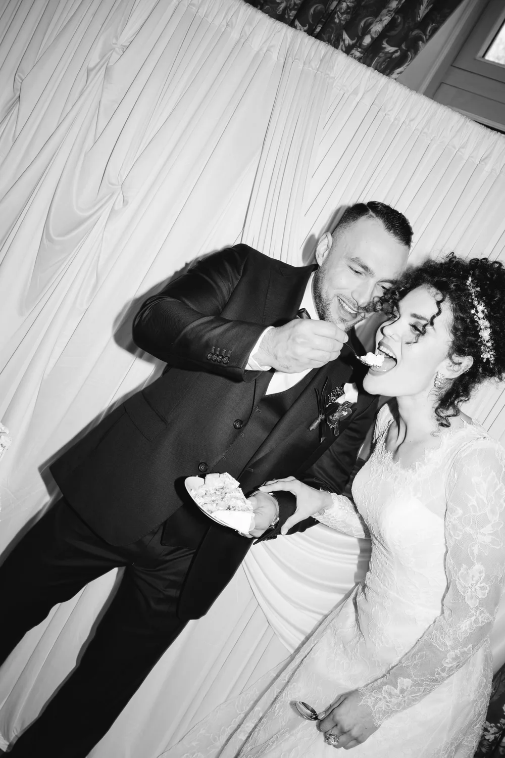 En noir et blanc, le marié fait goûter le gâteau à la mariée, devant un grand drapé blanc ; tous deux rient
