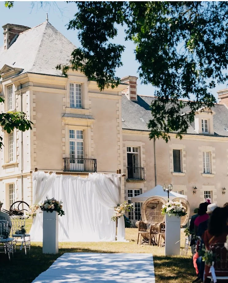 Le Château de Cop-Choux, façade de pierre claire, et sa cérémonie en plein air : fond de voilages blancs, colonnes de fleurs blanches, fauteuils paon en rotin et allée blanche