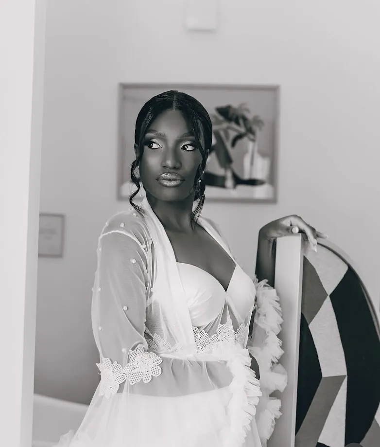 En noir et blanc, la mariée, dans un peignoir transparent orné de perles, regarde au loin dans sa chambre