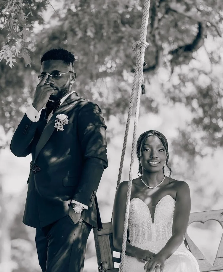 En noir et blanc, le marié debout, la main sur ses lunettes, et la mariée assise sur une balançoire en corde, sous de grands arbres