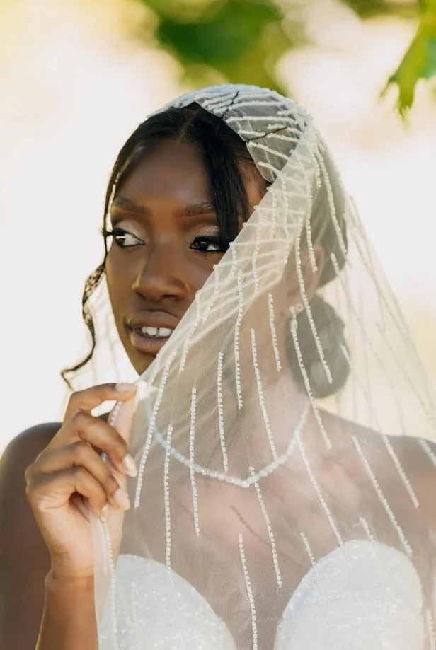 Portrait de la mariée, le regard au loin, une main sur son voile brodé de lignes de perles