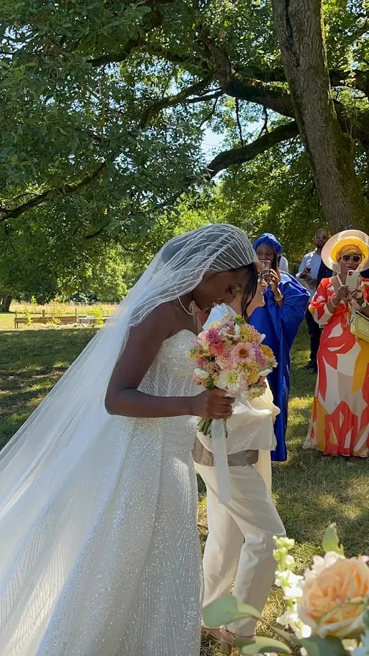 La mariée, un bouquet de fleurs pastel dans les mains, son voile derrière elle, sort sous les arbres, entourée d’invités en tenues colorées