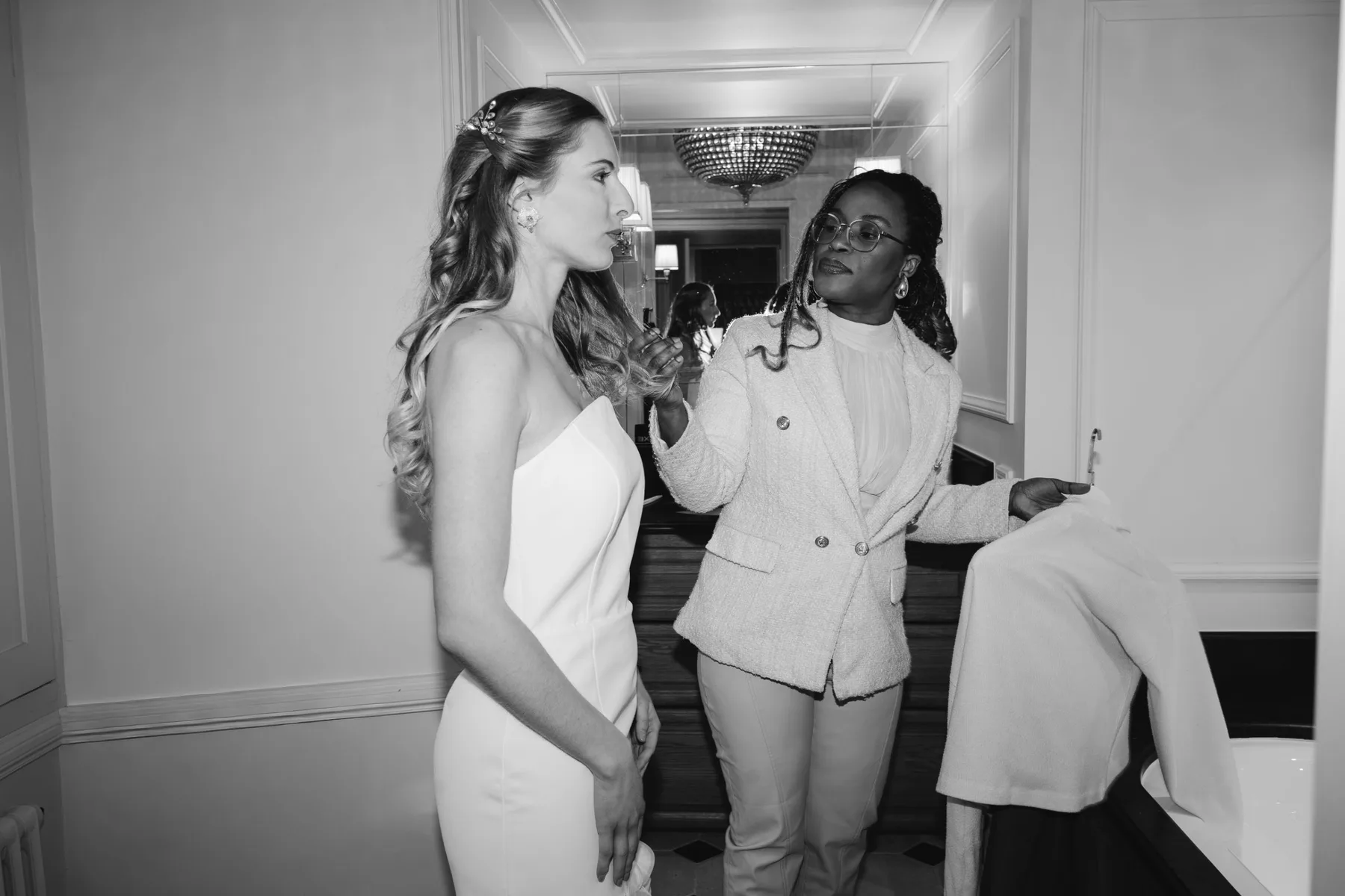 En noir et blanc, dans une salle de bains de château, une femme en blazer clair ajuste la coiffure de la mariée en robe bustier, un vêtement sur le bras