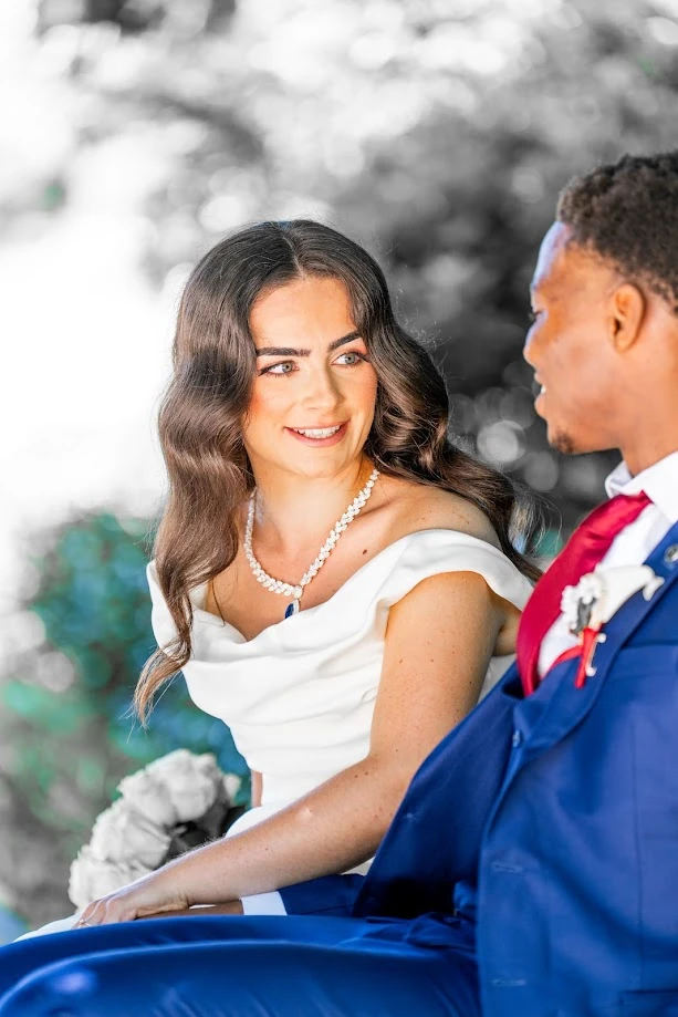 Portrait des mariés : la mariée aux cheveux bruns ondulés, un collier de perles sur sa robe blanche, sourit en regardant le marié, en costume bleu et cravate rouge