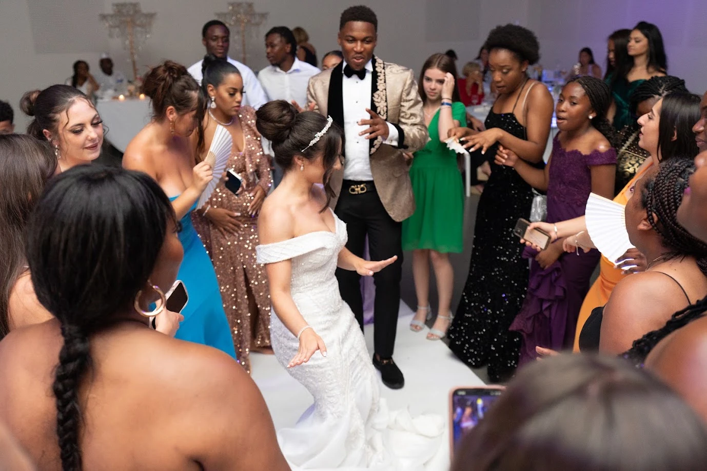 Sur la piste de danse, la mariée en robe blanche danse au milieu des invités en tenues colorées, le marié en veste dorée à ses côtés, des chandeliers de cristal en arrière-plan