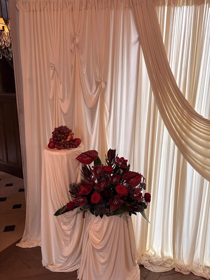 Fond de voilages crème et grande composition de fleurs rouges et bordeaux posée sur un piédestal drapé, avec des raisins en coupe sur un second piédestal
