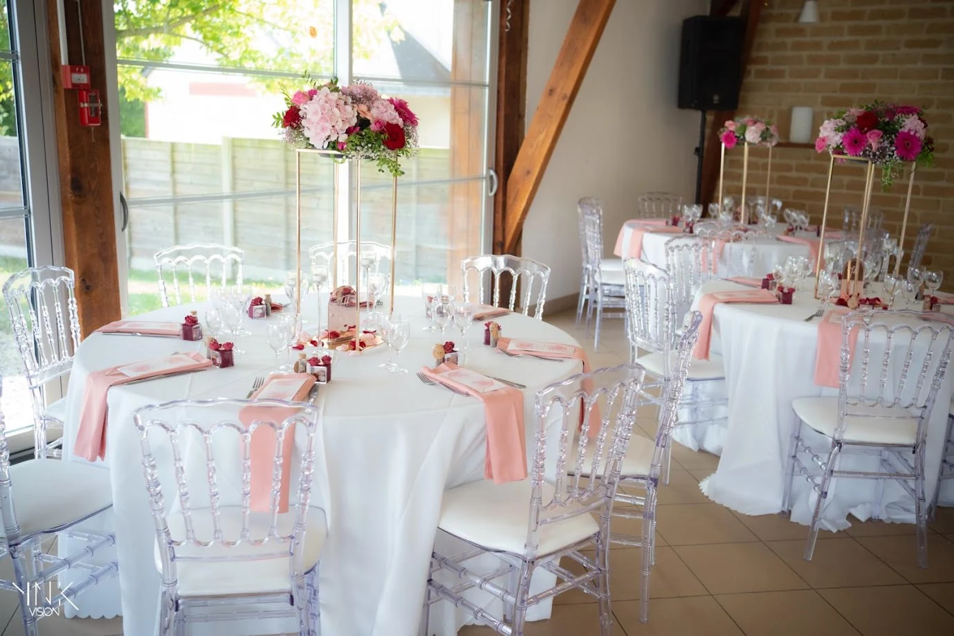 Salle aux poutres de bois et aux grandes baies vitrées : tables rondes aux chaises transparentes, serviettes roses et hauts centres de table de fleurs roses et fuchsia sur des socles dorés