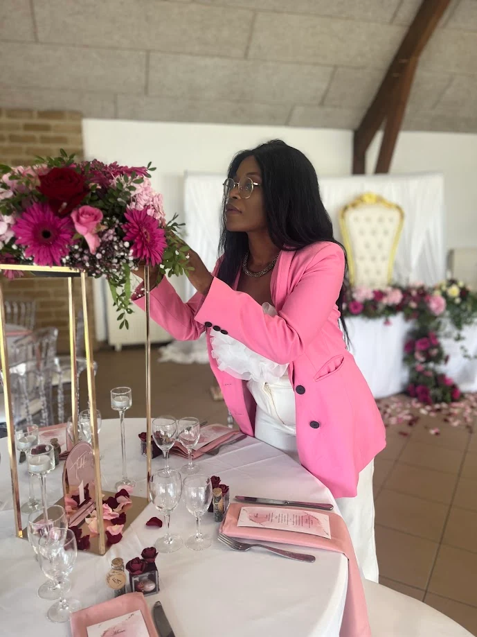La wedding planner, en blazer rose, ajuste un haut centre de table de gerberas et de roses roses sur une table ronde aux verres et aux pétales