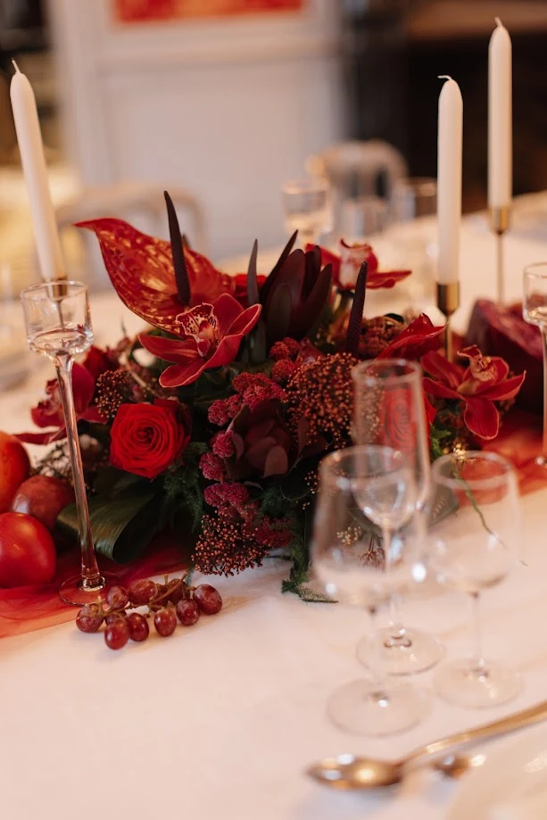 Centre de table de fleurs rouges et bordeaux, anthuriums, orchidées et roses, entre des bougies blanches et des verres sur une nappe blanche