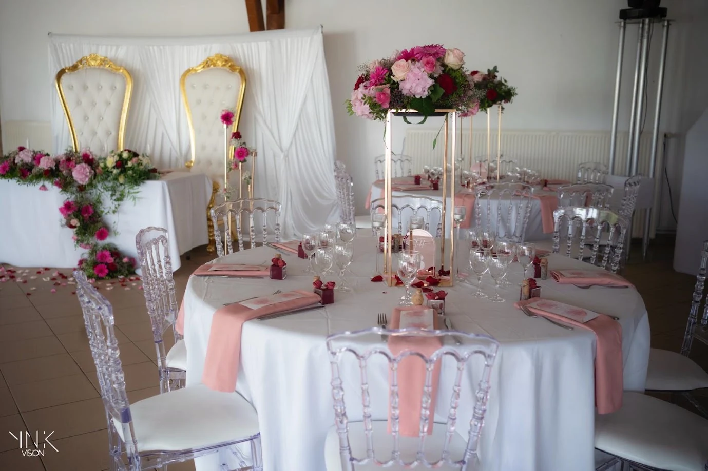 Table ronde dressée sous un haut centre de table de fleurs roses sur un socle doré, serviettes rose pâle, chaises transparentes ; au fond, la table d’honneur et ses fauteuils dorés