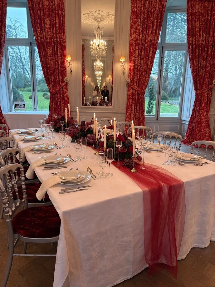 Une longue table dressée dans un salon de château aux rideaux rouges : chemin d’organza rouge, bougies, fleurs et lustre de cristal reflété dans le miroir