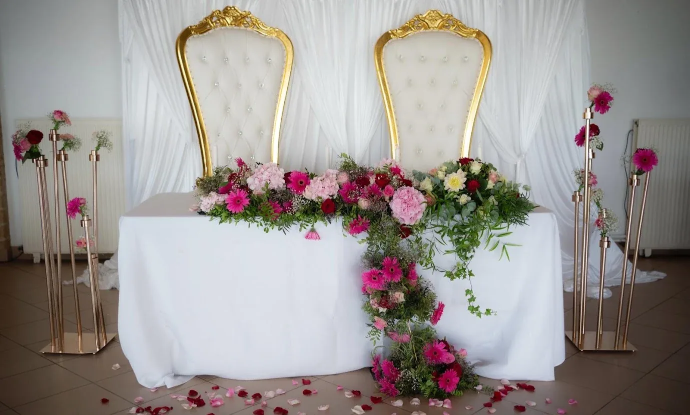 Table d’honneur : deux grands fauteuils blancs et dorés devant un fond de voilages blancs, une guirlande de fleurs roses, fuchsia et rouges qui retombe sur la table, des chandeliers dorés de chaque côté et des pétales sur le sol