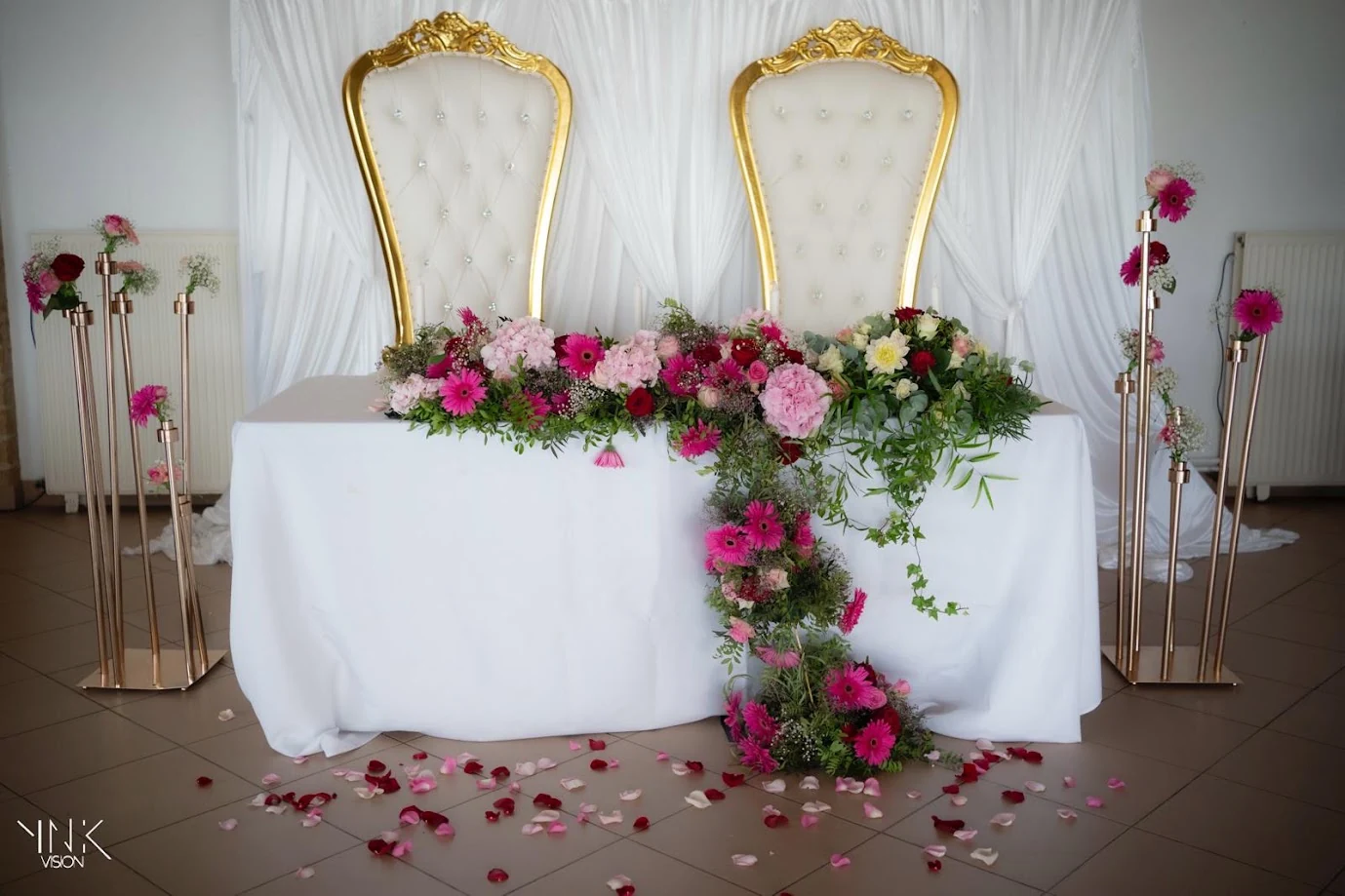 Table d’honneur : deux grands fauteuils blancs et dorés devant un fond de voilages blancs, une guirlande de fleurs roses, fuchsia et rouges qui retombe sur la table, des chandeliers dorés de chaque côté et des pétales sur le sol