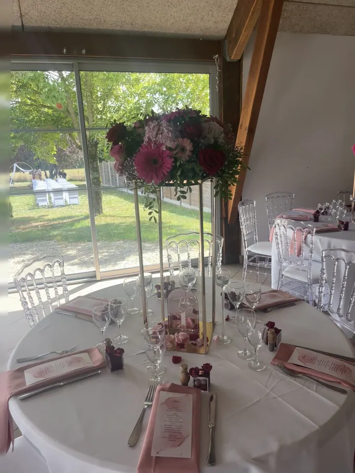Film : une table ronde dressée sous une verrière, avec un haut centre de table de fleurs roses et prunes, des menus imprimés et des chaises transparentes, le jardin en fond