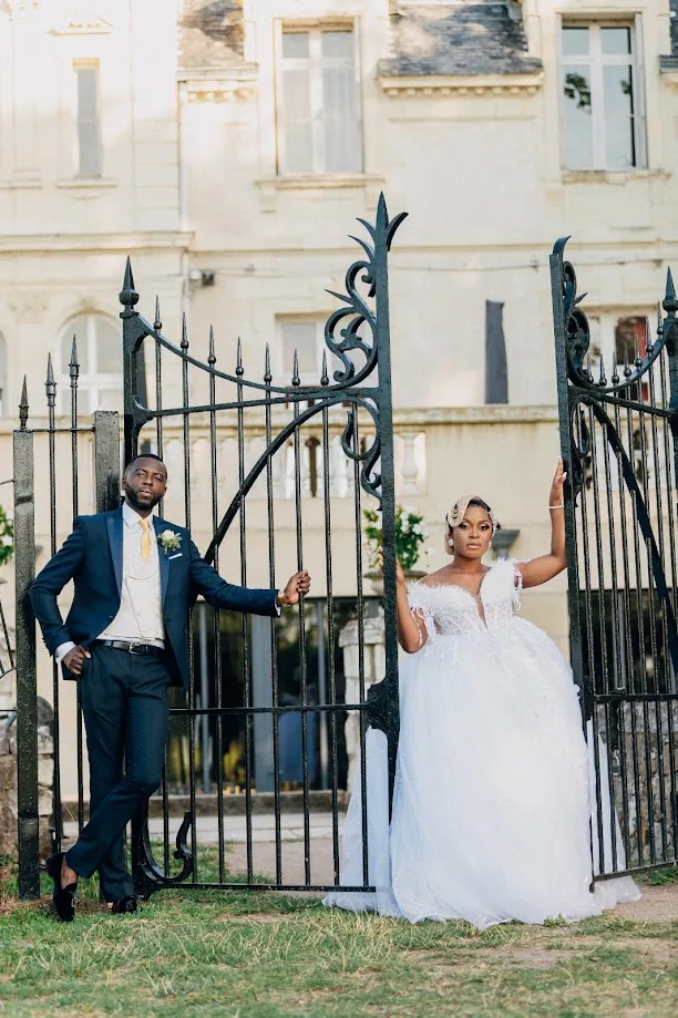 Devant la grille en fer forgé d’un château, le marié en costume bleu marine et la mariée en robe de tulle blanche posent de part et d’autre du portail