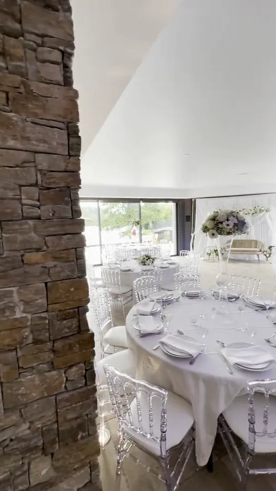 Film : une salle aux grandes baies vitrées, des tables rondes aux chaises transparentes et une longue table ornée de vases de fleurs blanches, sous un éclairage violet au plafond