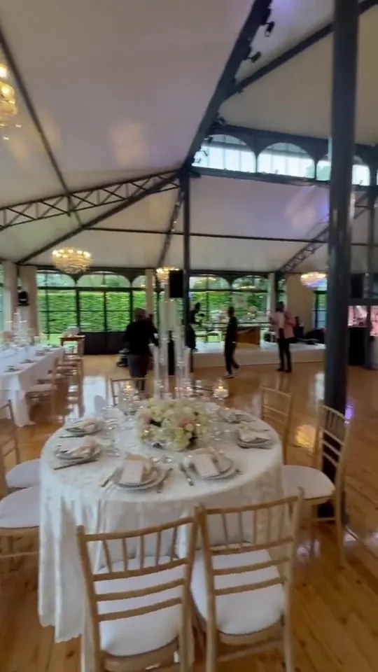 Film : une salle sous une verrière aux armatures métalliques, un lustre de cristal, des tables rondes dressées de fleurs blanches sur un parquet clair