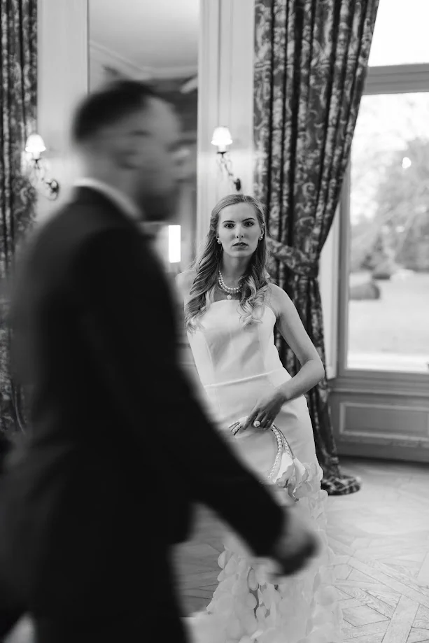 En noir et blanc, la mariée, une main sur la hanche, regarde l’objectif devant un rideau de brocart, tandis que le marié passe, flou, au premier plan