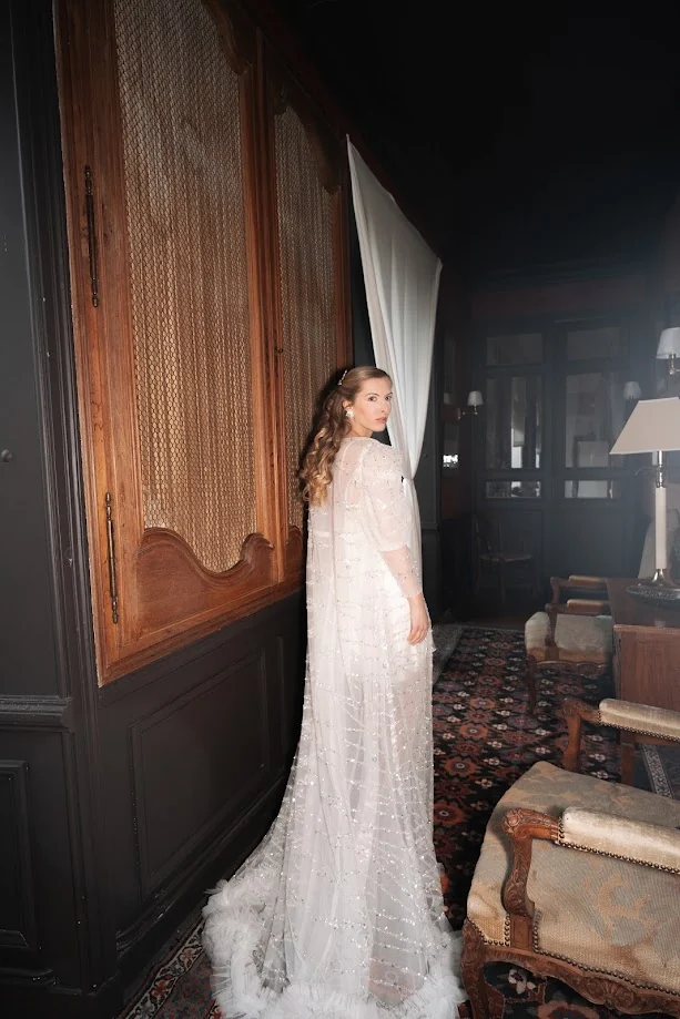 La mariée, de dos, tournée vers l’objectif, dans une robe de tulle brodé de perles et plumes, dans un salon aux boiseries sombres