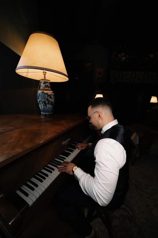 Le marié, en gilet noir et chemise blanche, joue du piano à la lumière d’une lampe de porcelaine bleue et blanche