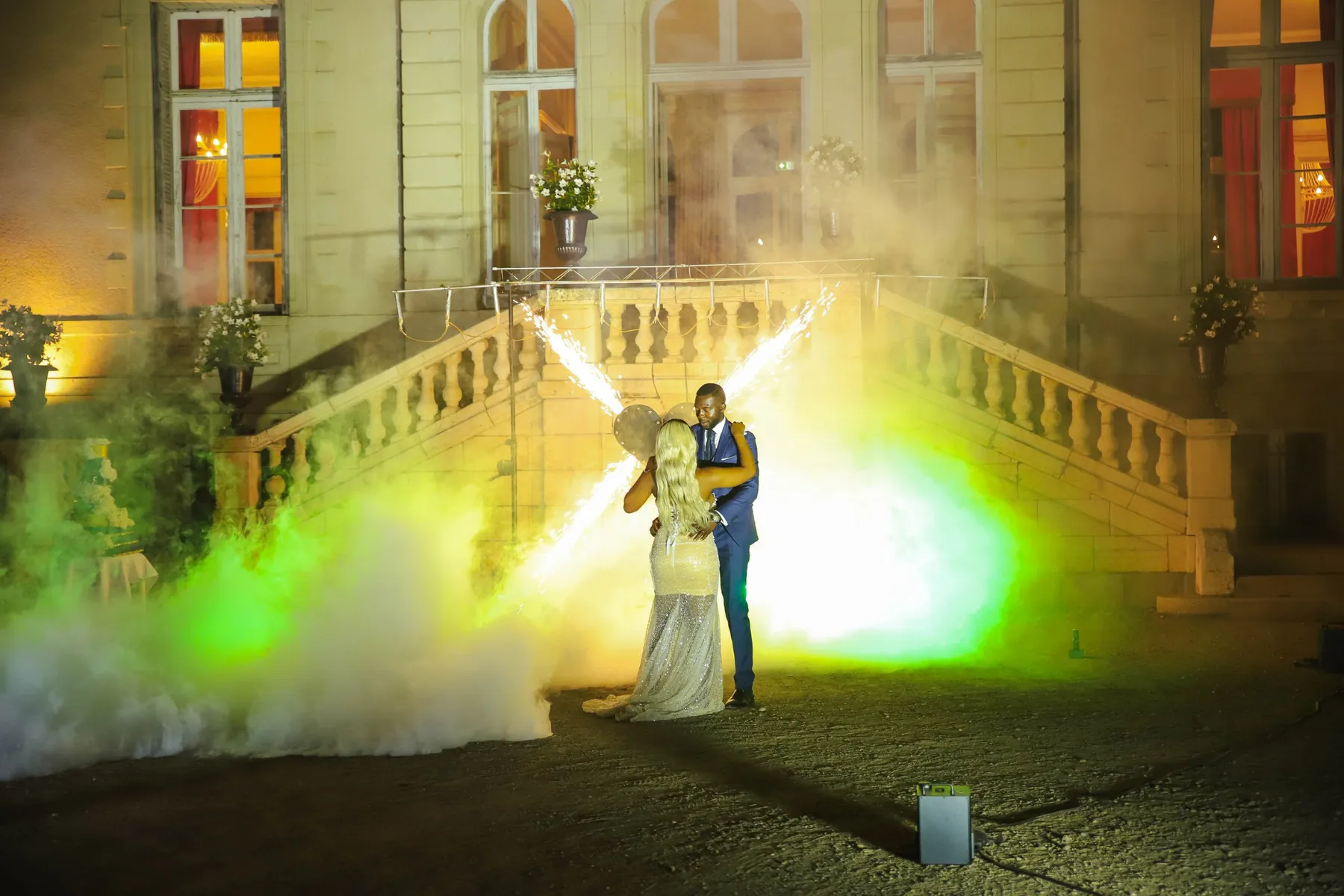 Sur le parvis d’un château, la nuit, les mariés dansent enlacés dans la fumée colorée, entre deux gerbes d’étincelles