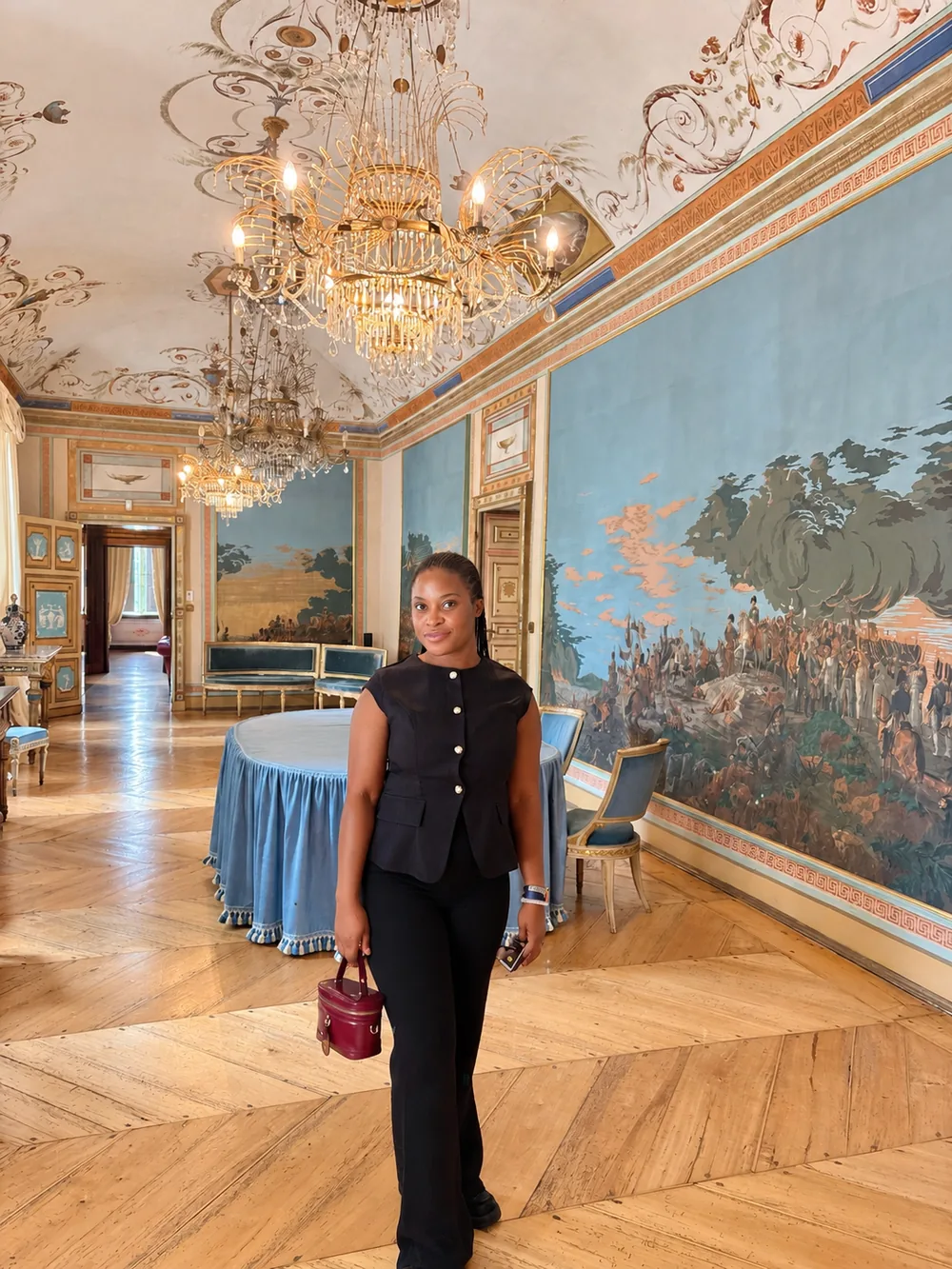 Gloire, en haut noir sans manches et pantalon noir, sourit dans un grand salon aux murs peints, aux lustres en cristal et au parquet ancien