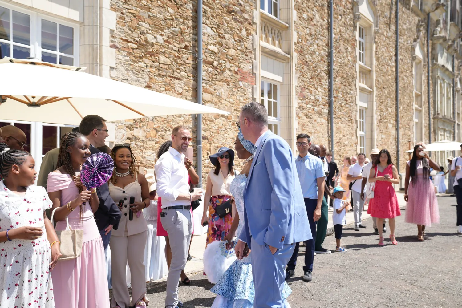 Devant le château, la mariée en tenue bleu ciel s’entretient avec le marié et des invités souriants, enfants et adultes, venus les saluer