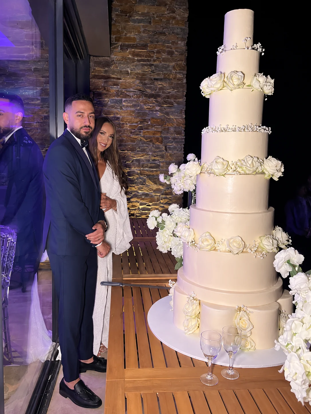 Hortense et Zoheir posent à côté d’une pièce montée de sept étages ornée de roses blanches et de gypsophile, sur une terrasse en bois la nuit
