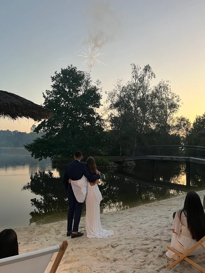Hortense et Zoheir, enlacés et vus de dos au bord d’un lac au crépuscule, regardent un feu d’artifice