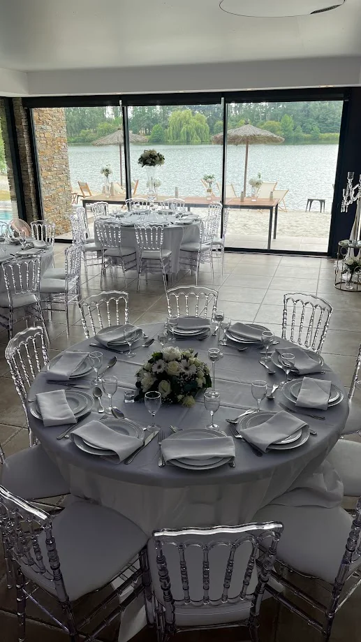 Une table ronde dressée, nappe grise, chaises transparentes et centre de table de roses blanches, face à une baie vitrée ouverte sur le lac