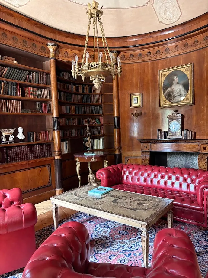 Une bibliothèque en bois sous une coupole peinte, avec un lustre doré, un portrait encadré et des canapés Chesterfield rouges