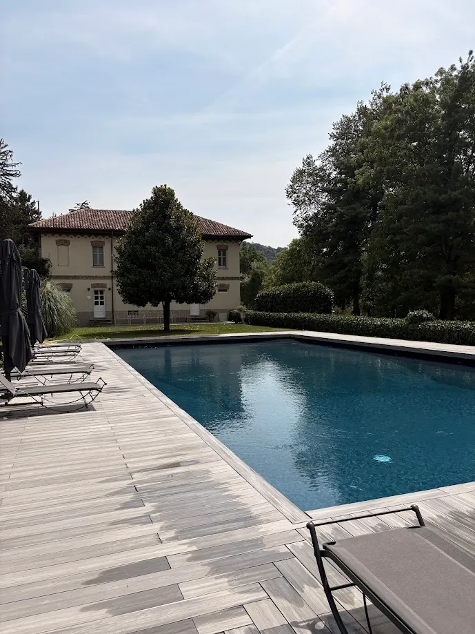 La piscine bordée d’un ponton de bois clair et de transats, devant une maison aux murs ocre et au toit de tuiles, entourée d’arbres