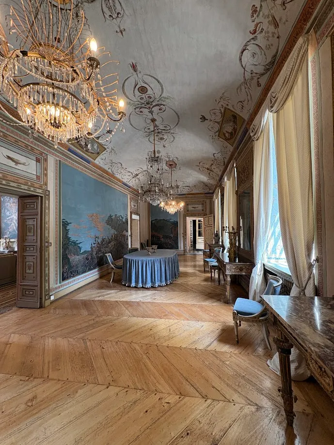 Un grand salon au plafond peint, éclairé de lustres en cristal, avec une table ronde à la nappe bleue et des fresques murales