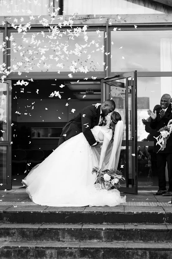 En noir et blanc, sur les marches d’une salle, le marié penche la mariée voilée pour l’embrasser sous une pluie de papillons blancs, des invités applaudissent à droite