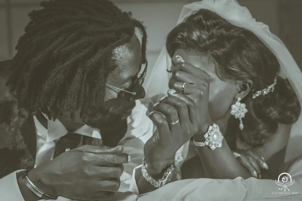 Dans une image tendre en noir et blanc, le marié, lunettes de soleil, se penche vers la mariée qui cache son visage dans ses mains, bagues et bijoux en évidence