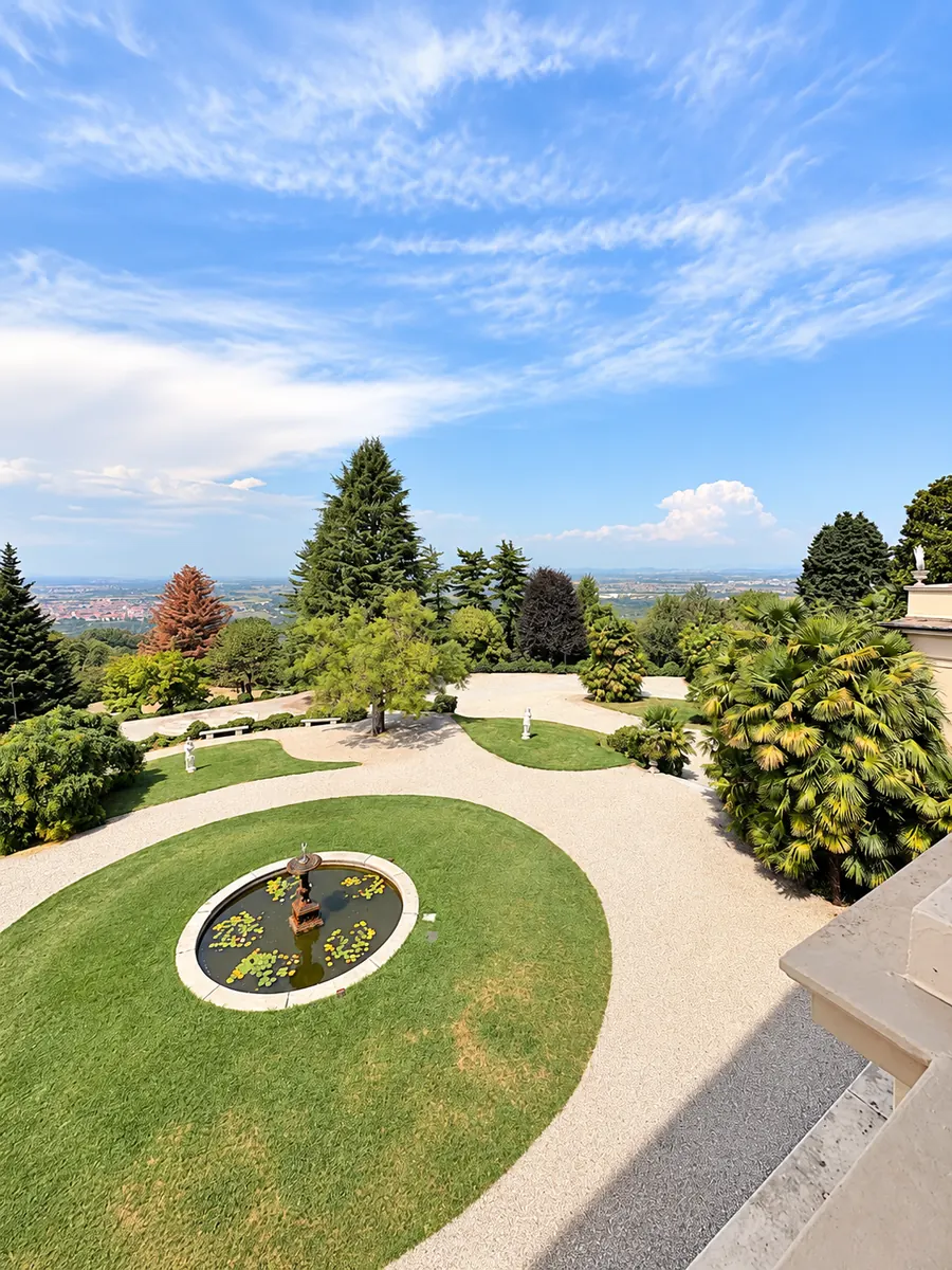 Vu d’en haut, un jardin à l’italienne : bassin circulaire au milieu d’une pelouse, allées de gravier clair, palmiers et pins, avec la plaine au loin sous un grand ciel bleu