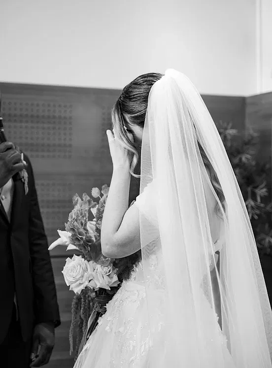 En noir et blanc, la mariée voilée porte la main à son visage, émue, près d’un homme qui tient un micro