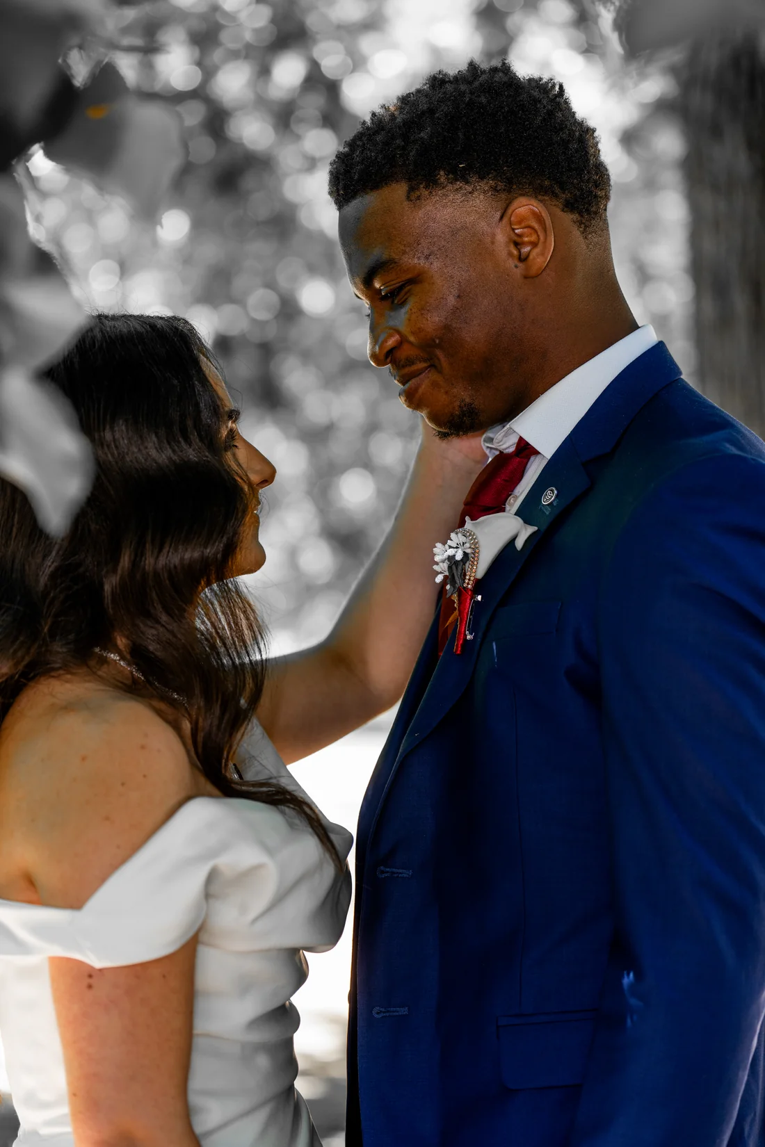 Un couple mixte se regarde tendrement : la mariée caresse le visage du marié, en costume bleu, cravate rouge