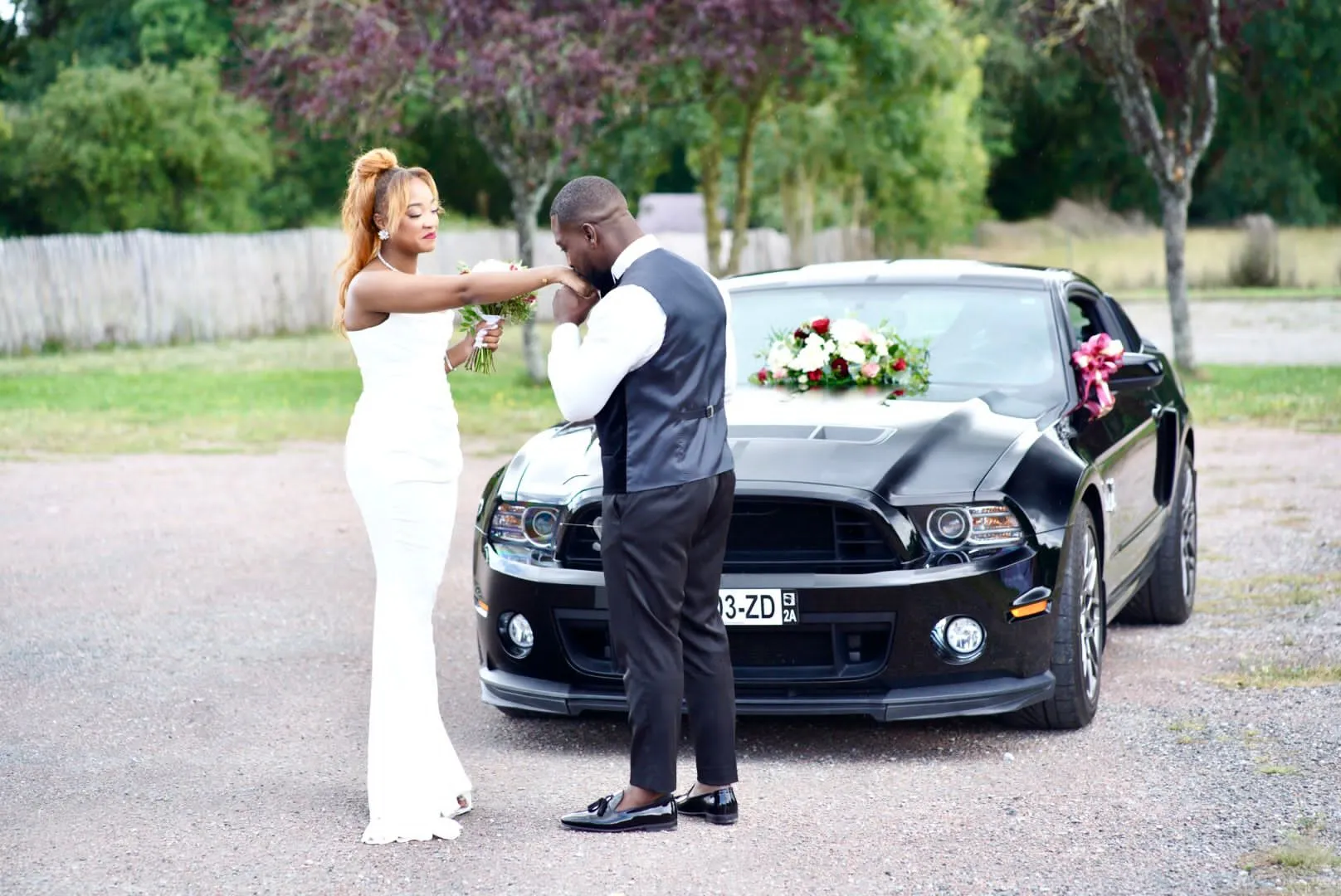 Devant une voiture noire ornée de fleurs, le marié baise la main de la mariée en robe blanche, bouquet à la main