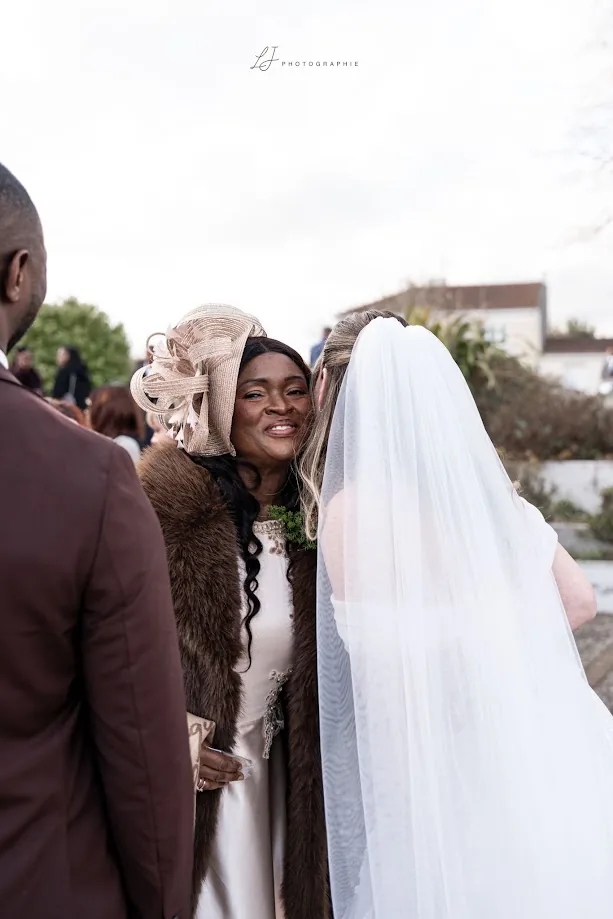 Une invitée au chapeau beige et au manteau de fourrure sourit en serrant la mariée, vue de dos sous son voile
