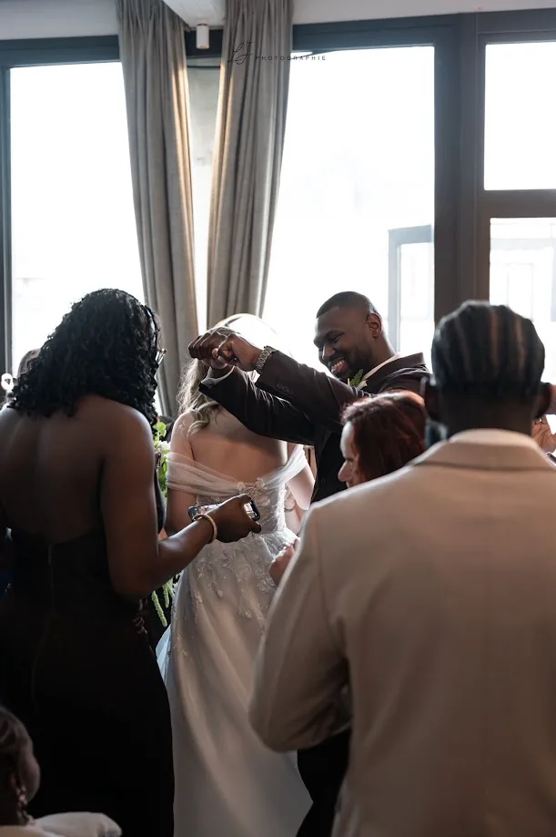 Près d’une grande baie vitrée, le marié, tout sourire, lève le poing en dansant avec la mariée vue de dos, entourés de leurs invités